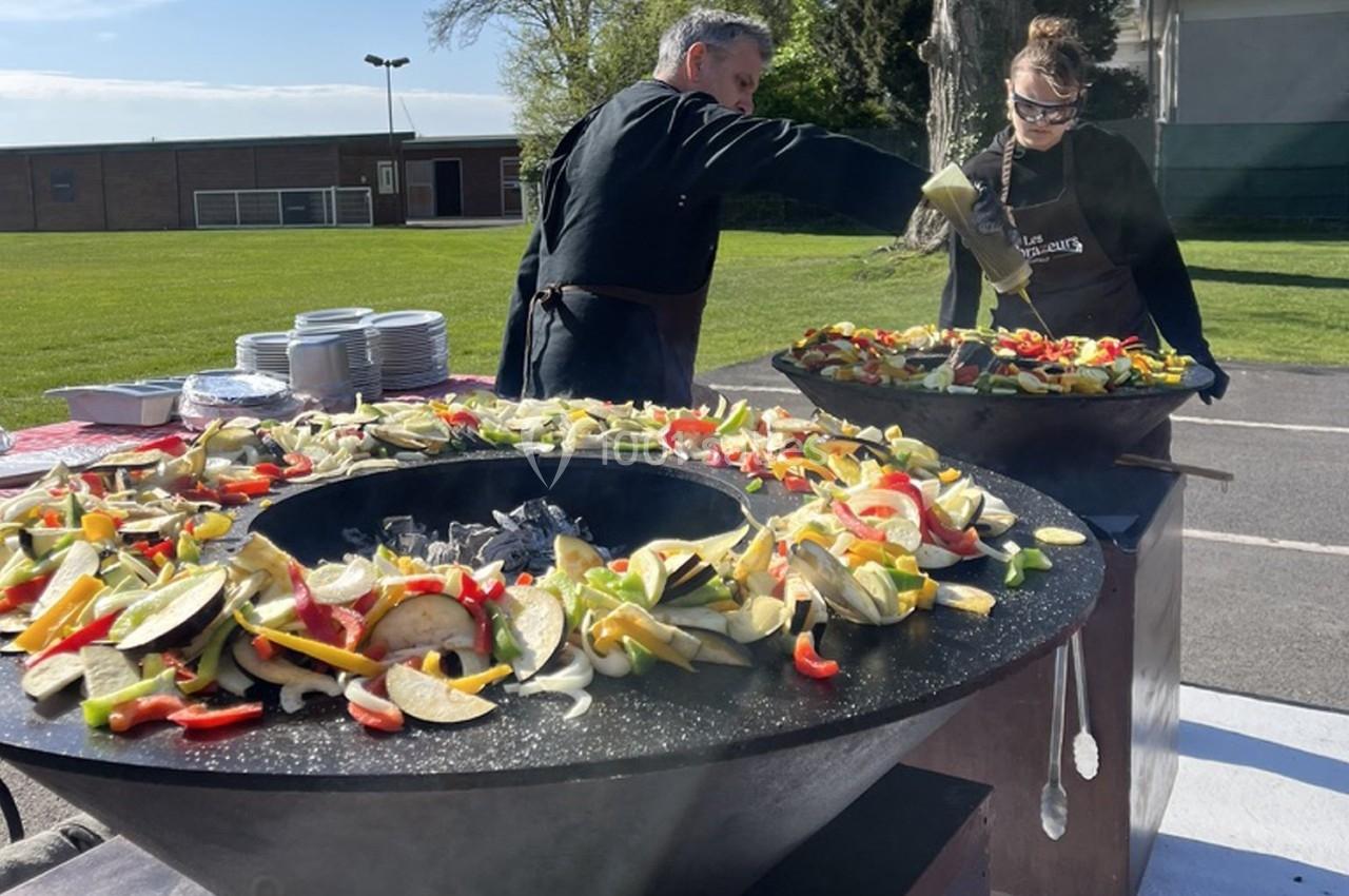 Deux personnes cuisinent des légumes variés sur de grandes plaques chauffantes en extérieur, près d'un espace vert.