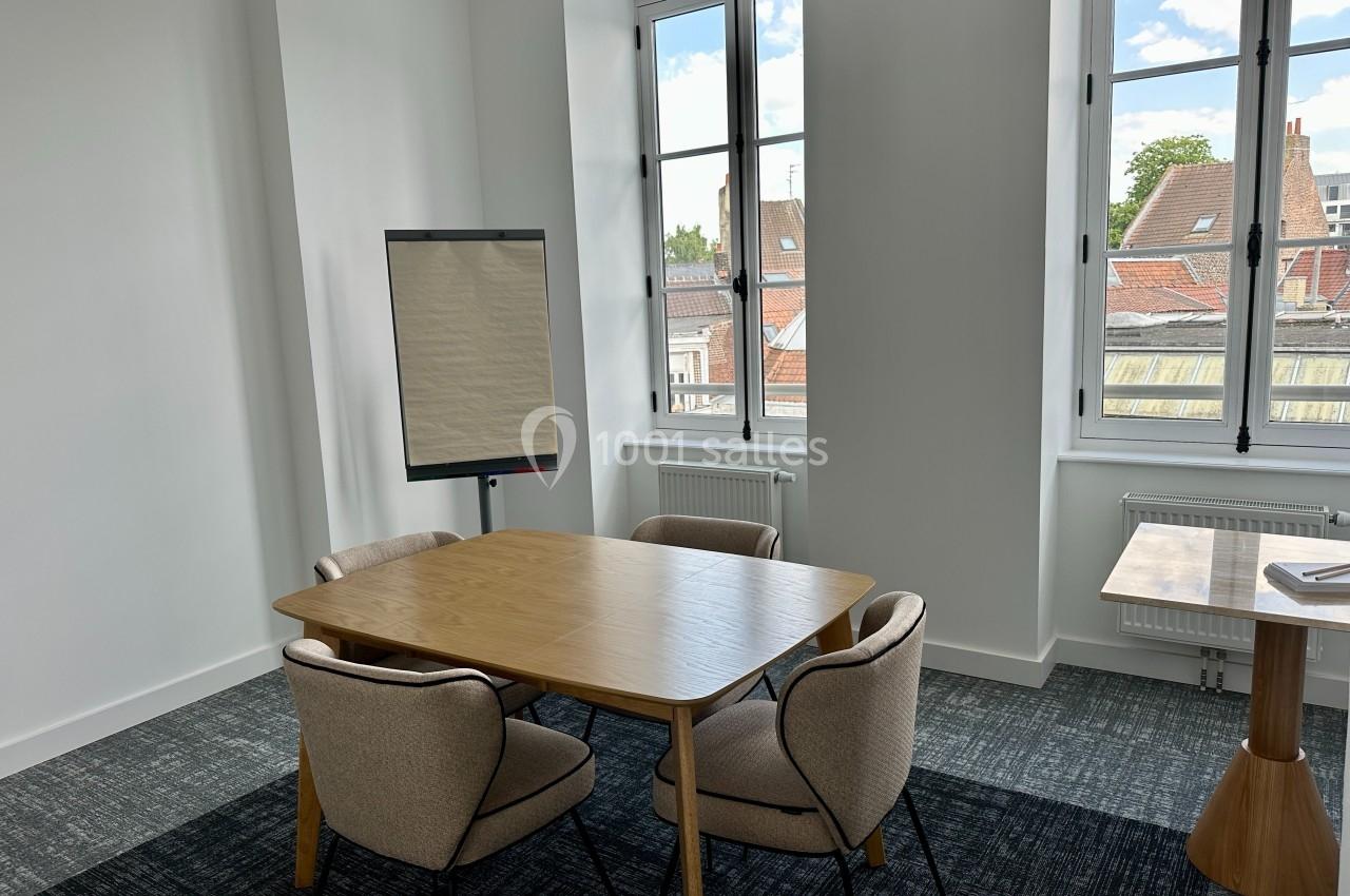 Salle de réunion lumineuse avec table en bois, quatre chaises, un paperboard et fenêtres donnant sur des toits.