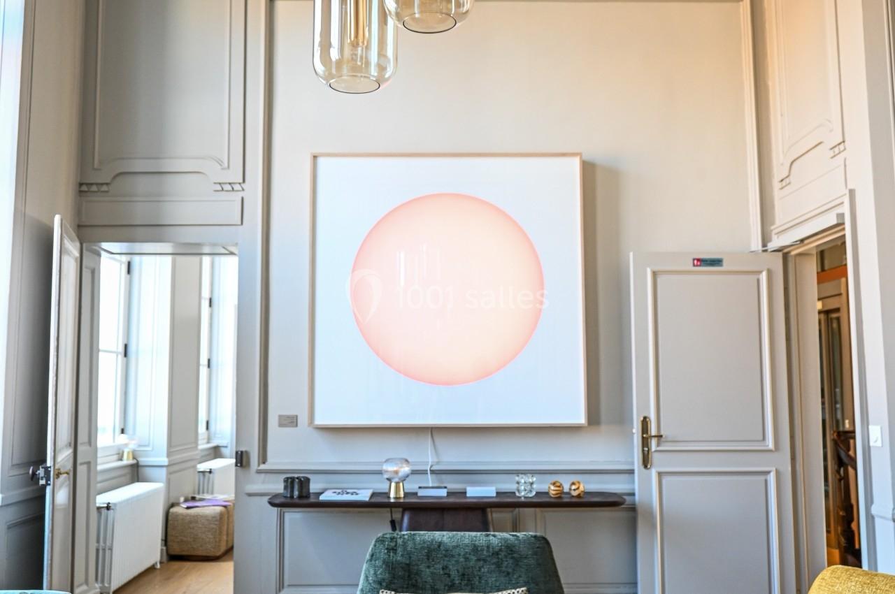 Salon lumineux avec fauteuils colorés, suspension moderne et tableau abstrait éclairé sur un mur beige.