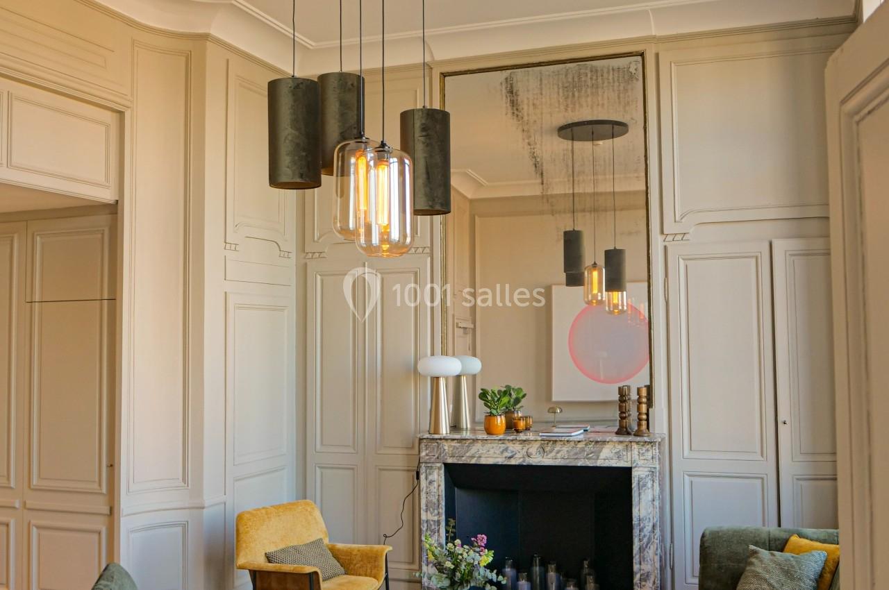 Salon élégant avec canapé vert, fauteuils jaunes, cheminée en marbre, miroir, et suspension lumineuse moderne.