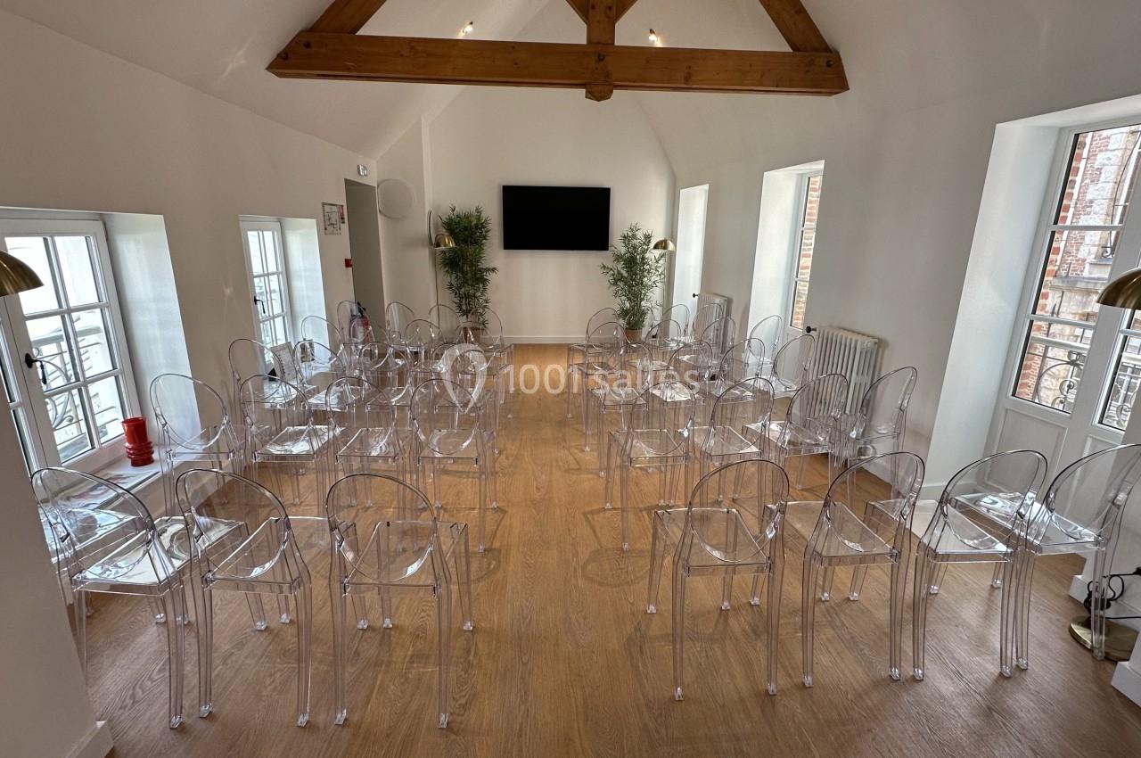 Salle lumineuse aménagée avec des chaises transparentes alignées, un écran mural et des poutres apparentes au plafond.