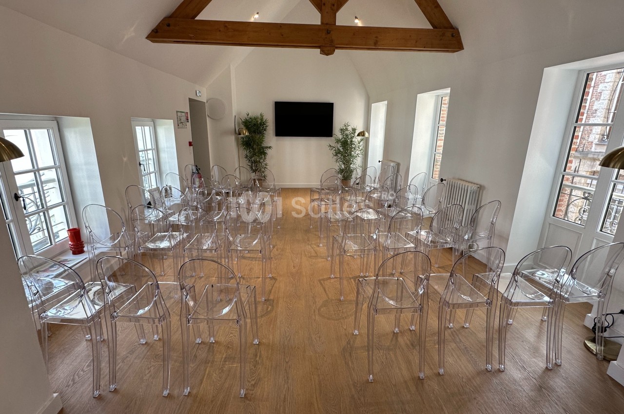 Salle lumineuse aménagée avec des chaises transparentes alignées, un écran mural et des poutres apparentes au plafond.