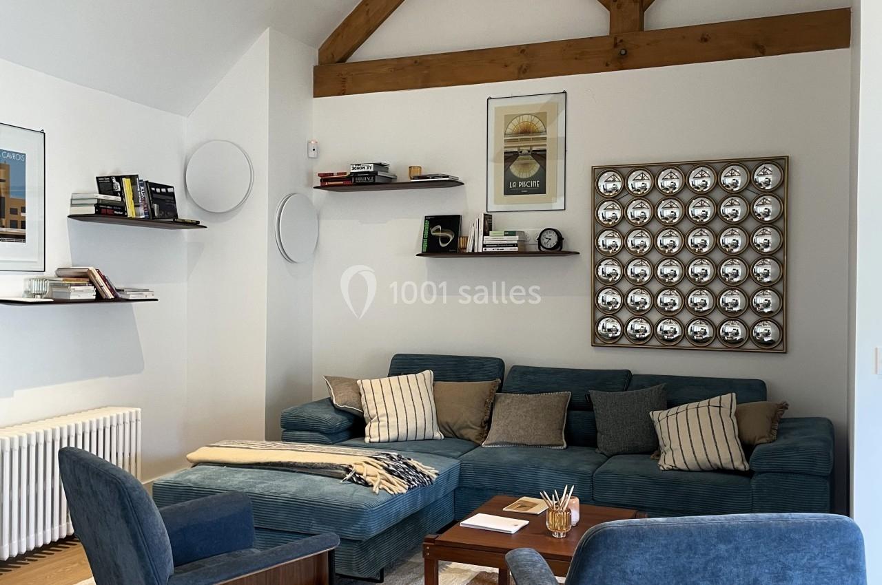 Salon moderne avec canapé bleu, fauteuils assortis, poutres apparentes, étagères murales et décoration murale géométrique.