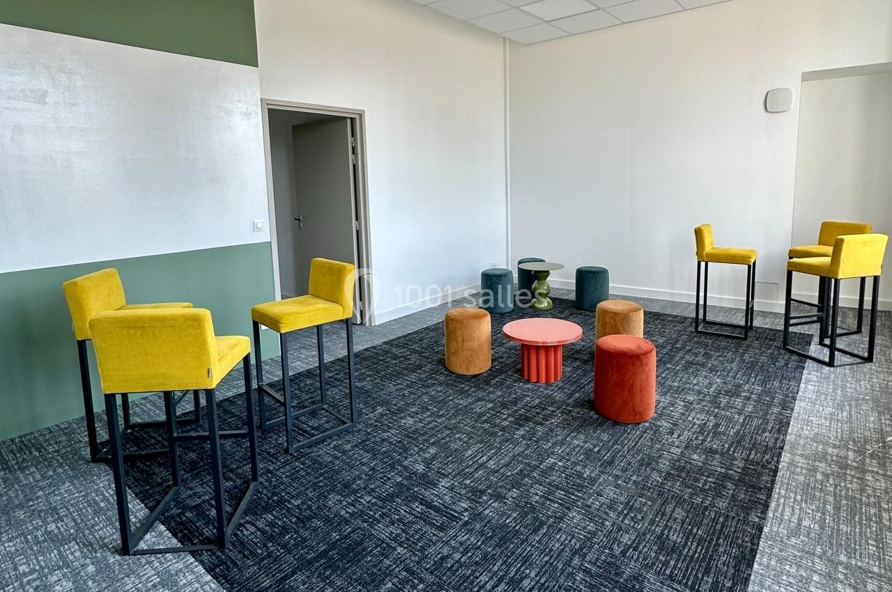Salle lumineuse avec chaises hautes jaunes, poufs colorés, table basse rouge et sol en moquette grise.