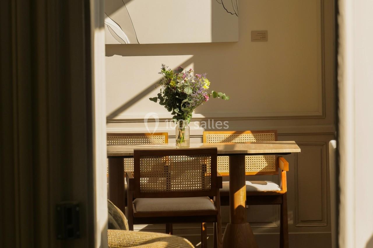Salle à manger lumineuse avec table en bois, chaises en rotin, bouquet de fleurs et tableau minimaliste au mur.