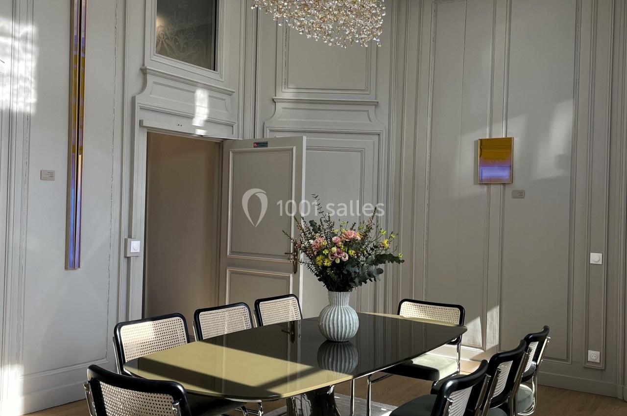 Salle à manger lumineuse avec une table en verre, des chaises modernes, un vase de fleurs et un lustre design.