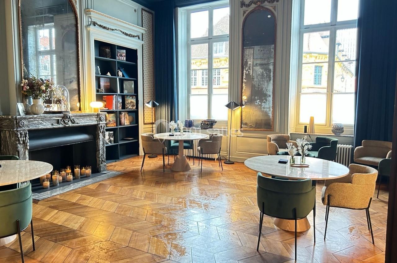 Salle élégante avec parquet en bois, tables rondes, fauteuils et cheminée décorée, éclairée par de grandes fenêtres.