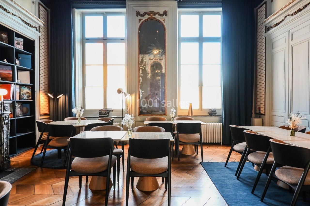 Salle lumineuse avec grandes fenêtres, tables en bois, chaises rembourrées et décorations murales élégantes.