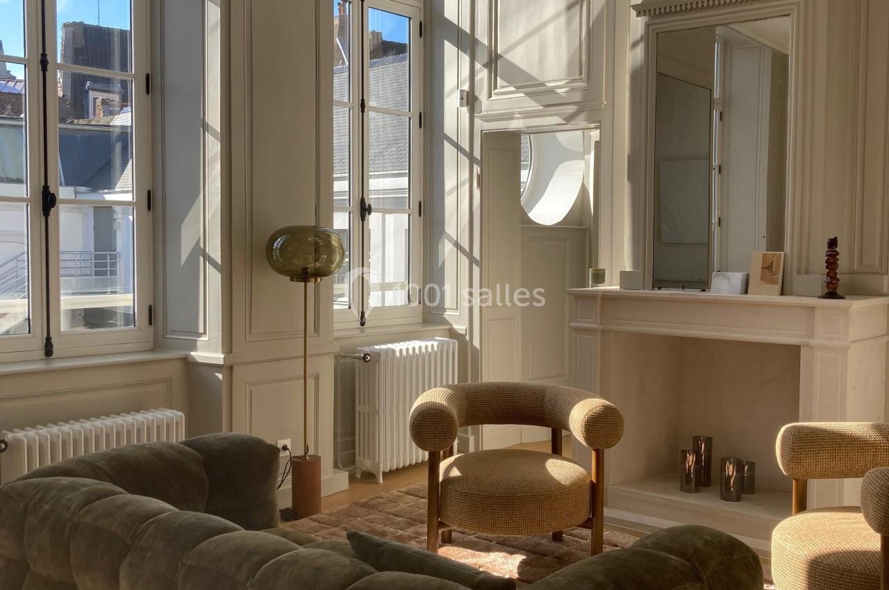 Salon lumineux avec canapé en velours, fauteuils, cheminée décorative et grandes fenêtres laissant entrer la lumière…