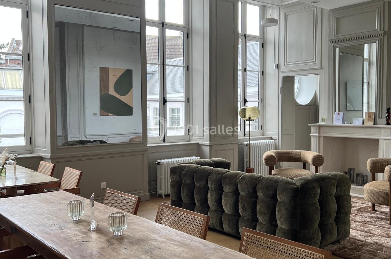 Salle lumineuse avec grandes fenêtres, table en bois, chaises, canapé vert, fauteuils beiges et décoration minimaliste.