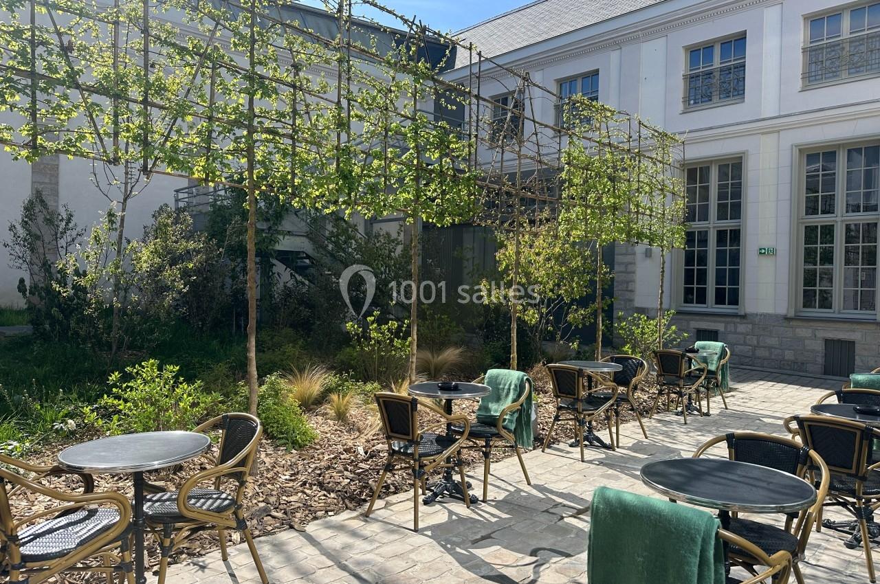 Terrasse ensoleillée avec tables et chaises en rotin, entourée de verdure et située devant un bâtiment blanc.
