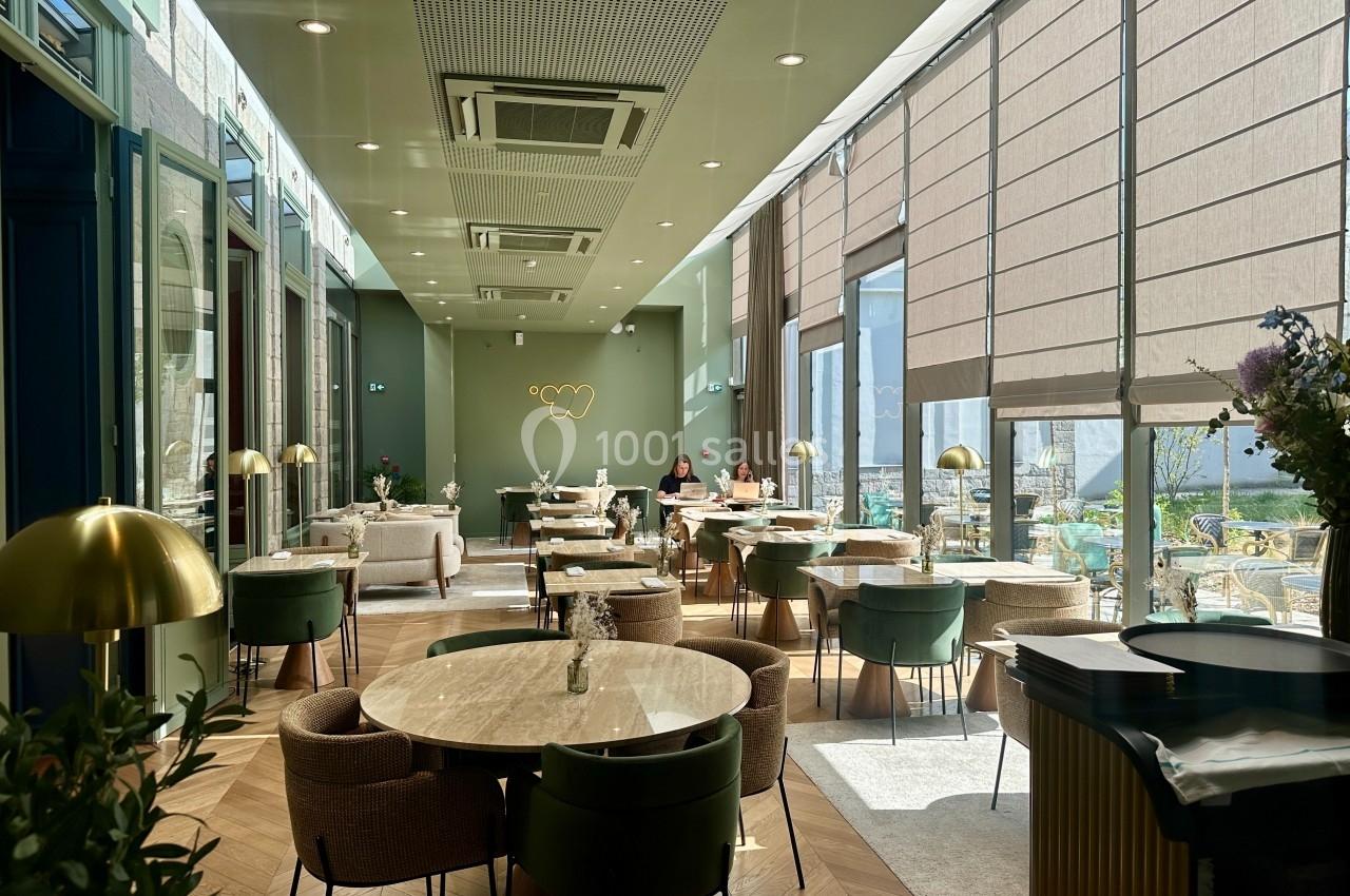 Salle de restaurant lumineuse avec grandes baies vitrées, tables en marbre, chaises vertes et beige, et décoration moderne.