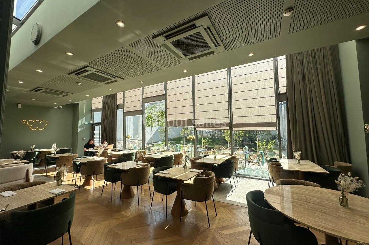 Salle lumineuse avec grandes baies vitrées, tables en bois clair, chaises modernes et vue sur un jardin extérieur.