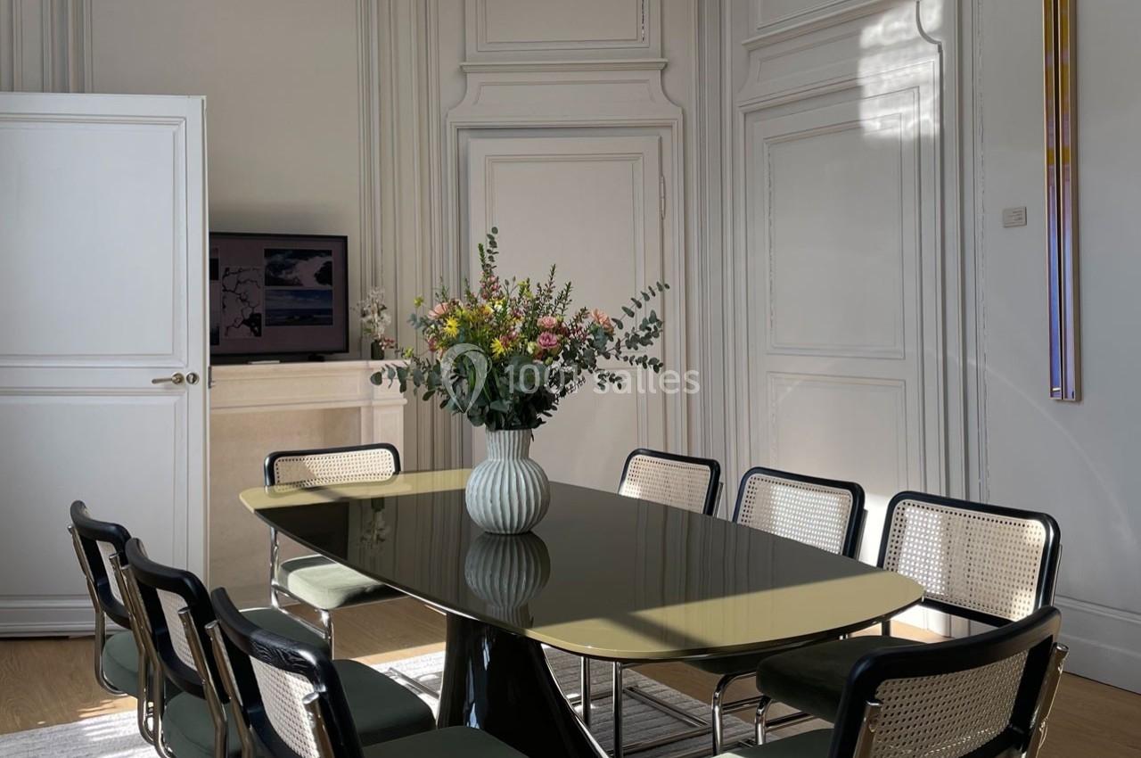 Salle à manger lumineuse avec une table ovale, des chaises en osier et un vase de fleurs colorées au centre.