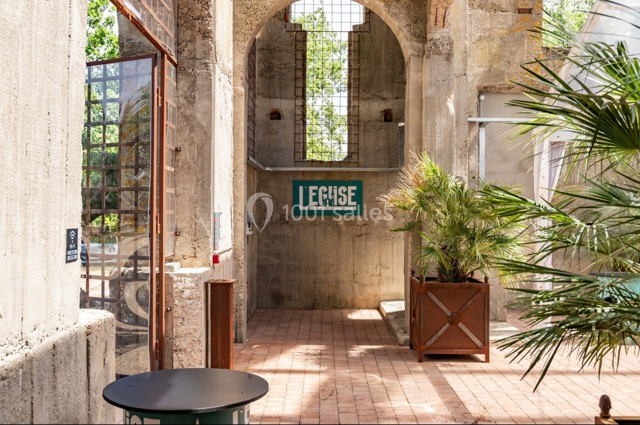 Entrée d'un bâtiment en béton avec une enseigne ’Le Cèdre’, des plantes en pot et des murs ouverts sur l'extérieur.