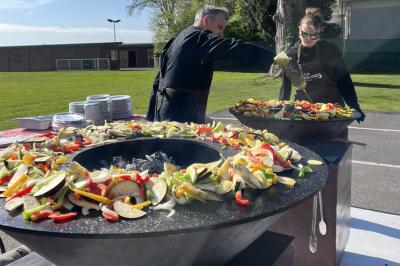 Deux personnes cuisinent des légumes colorés sur une grande plancha en extérieur, par une journée ensoleillée.