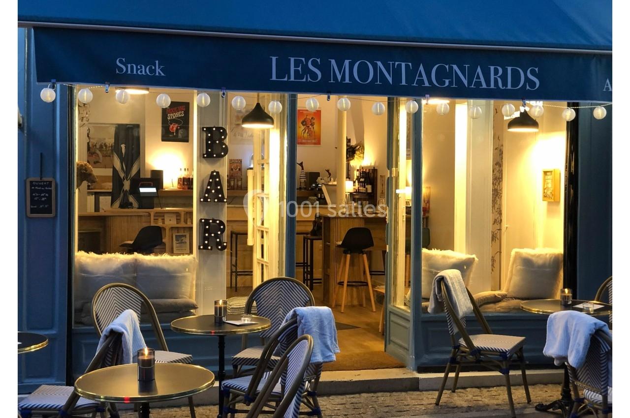 Façade d'un café-restaurant avec terrasse extérieure, tables rondes et enseigne lumineuse ’BAR’ visible à l'intérieur.