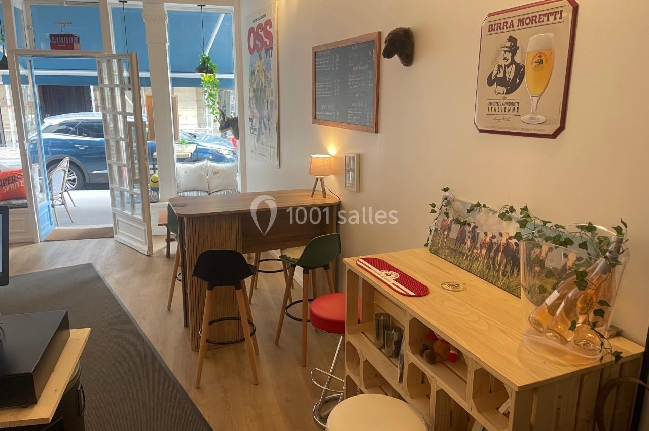 Intérieur d'un café avec tables en bois, tabourets, affiches décoratives et lumière naturelle provenant de la vitrine.