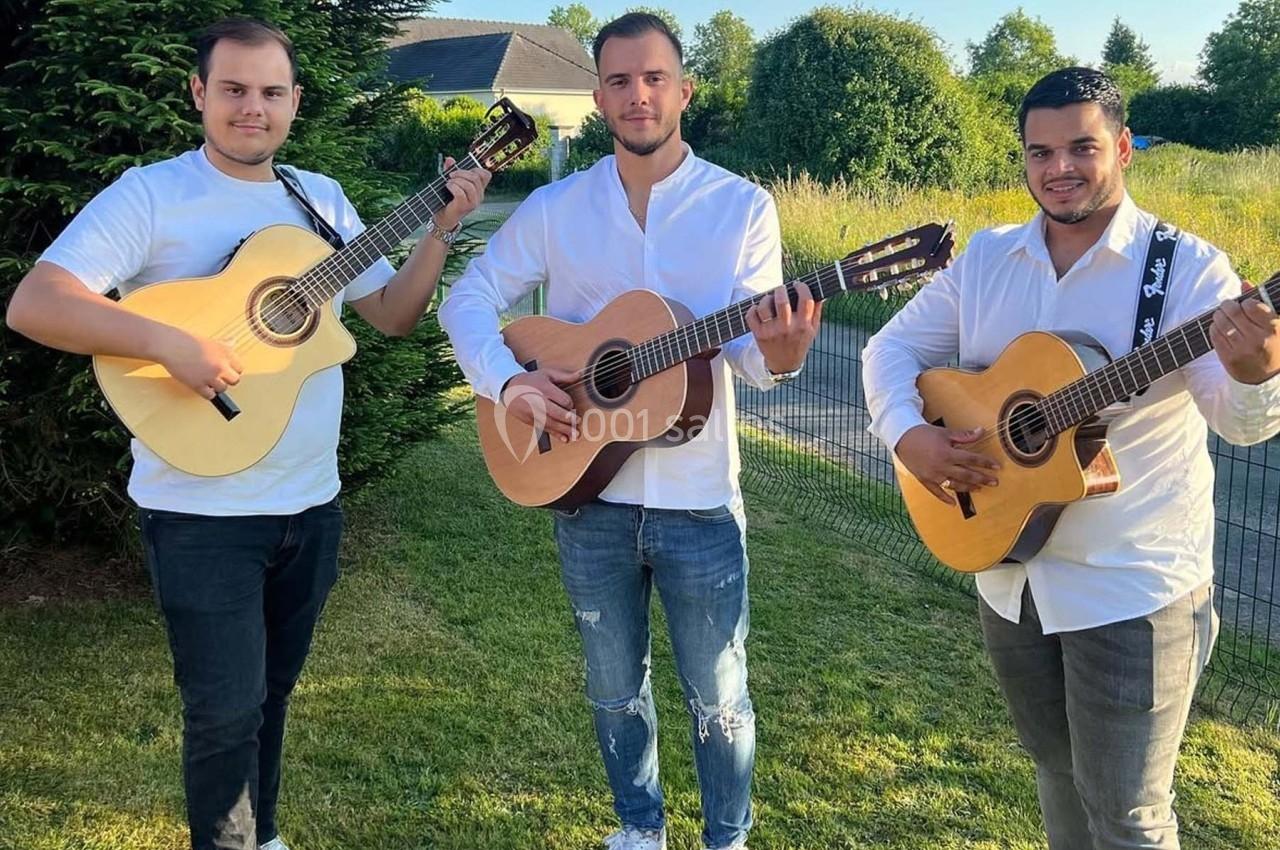 Trois hommes jouent de la guitare acoustique dans un jardin, vêtus de chemises blanches et entourés de verdure.