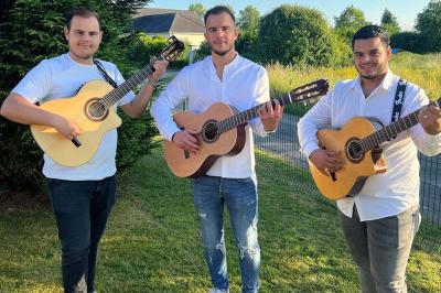 Trois hommes debout dans un jardin jouent de la guitare acoustique, vêtus de chemises blanches.