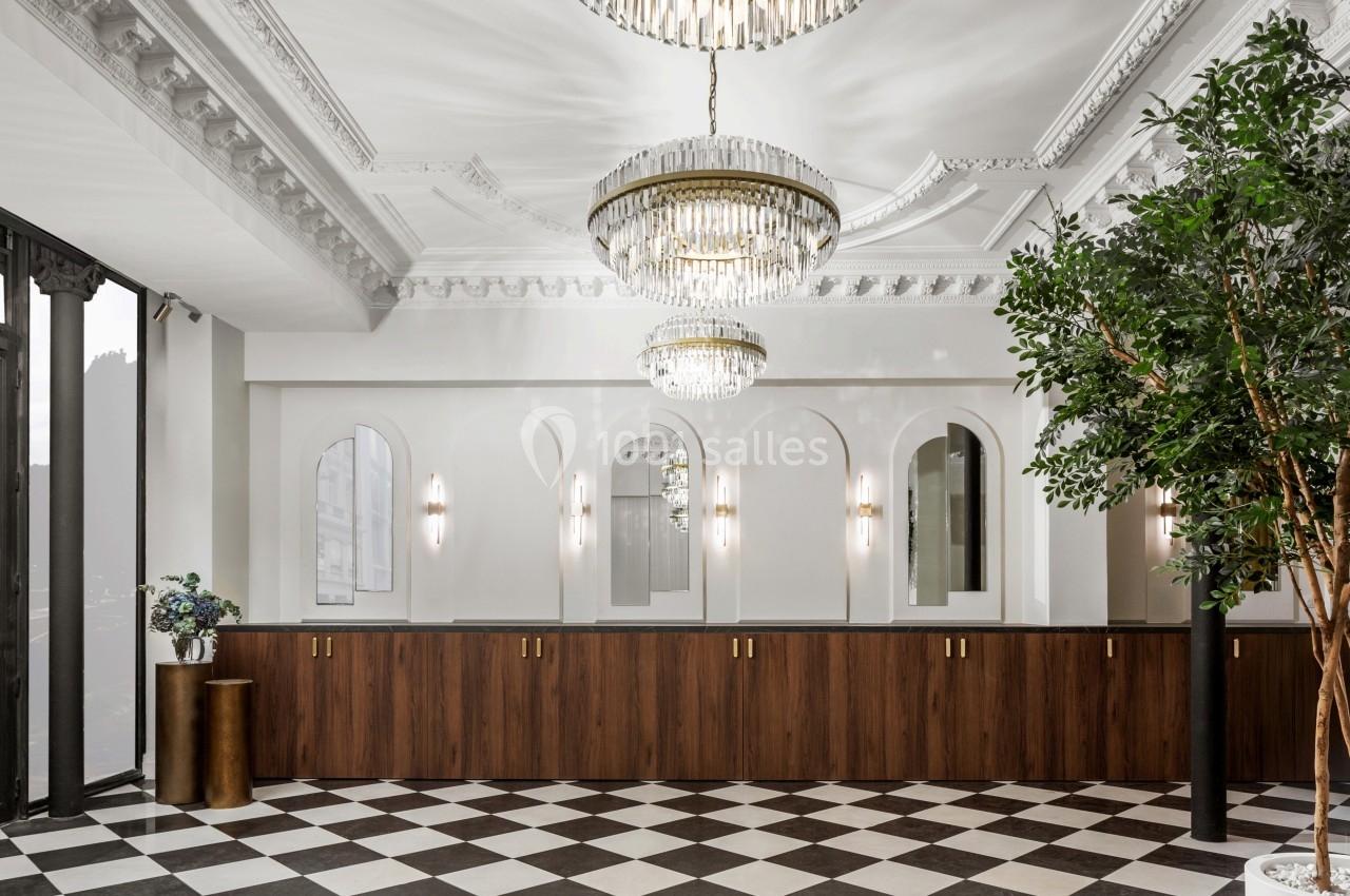 Hall élégant avec sol en damier, comptoir en bois, lustres en cristal, murs blancs et grandes fenêtres.