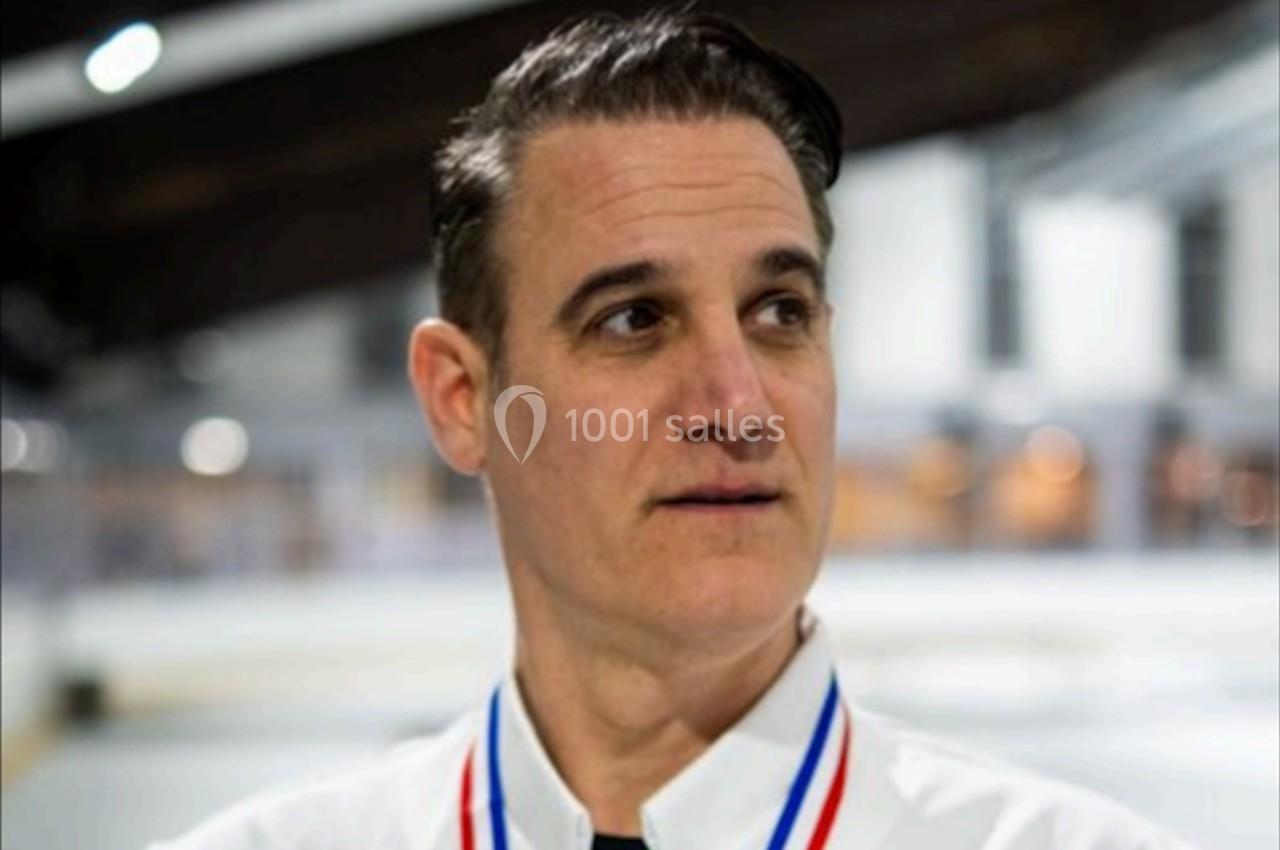 Homme portant une chemise blanche et une médaille, debout dans un environnement intérieur lumineux flou.