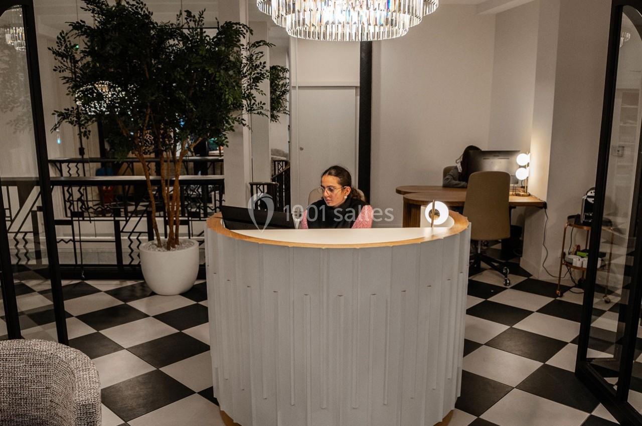 Femme assise à un comptoir d'accueil arrondi dans un espace intérieur moderne avec sol en damier et éclairage suspendu.
