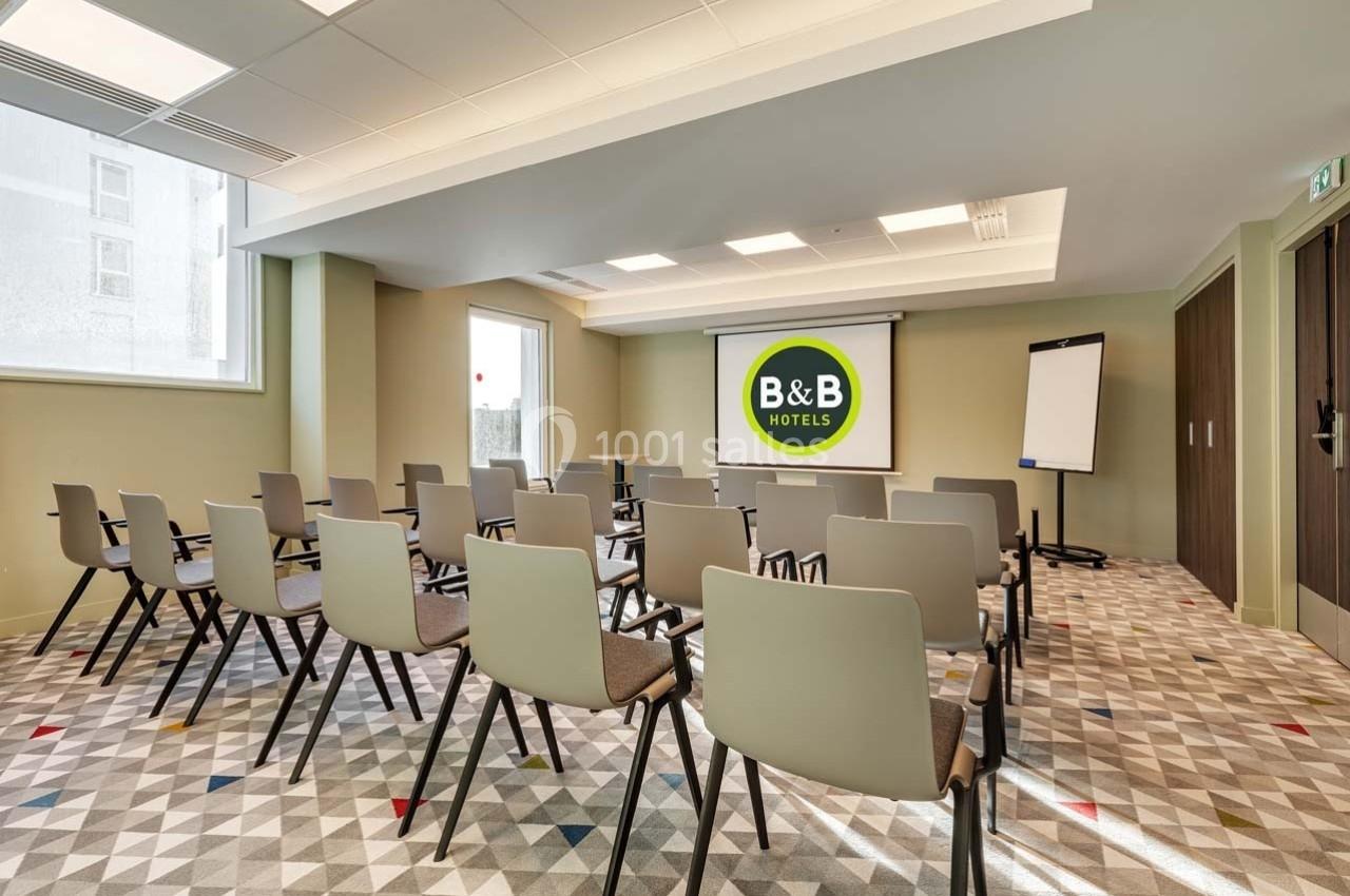 Salle de réunion lumineuse avec chaises alignées, écran de projection affichant le logo B&B Hotels et un tableau blanc.