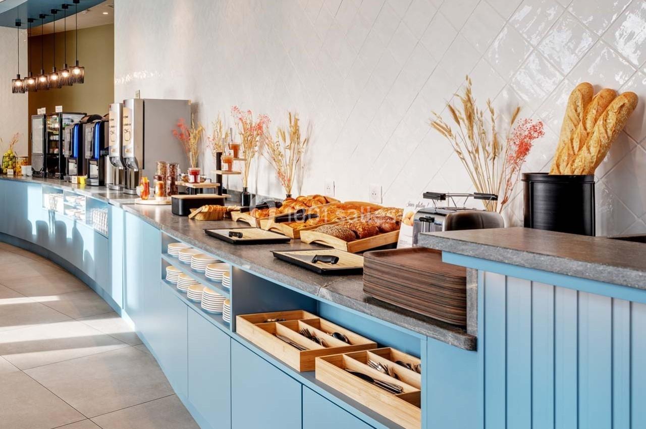 Buffet de petit-déjeuner avec viennoiseries, pains, boissons chaudes et couverts disposés sur un comptoir bleu.