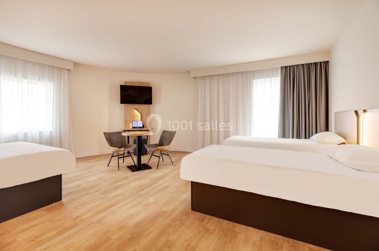 Chambre lumineuse avec deux lits simples, un coin table avec chaises, parquet clair et grandes fenêtres avec rideaux.