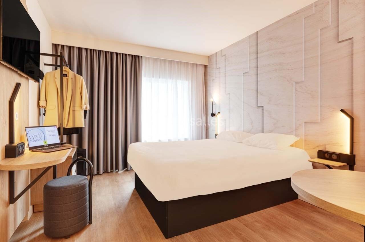Chambre d'hôtel lumineuse avec lit double, bureau équipé, fauteuil, rideaux opaques et éclairage moderne.