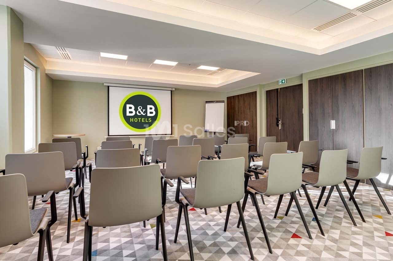 Salle de réunion lumineuse avec chaises alignées, écran de projection affichant le logo B&B Hotels et tableau blanc.