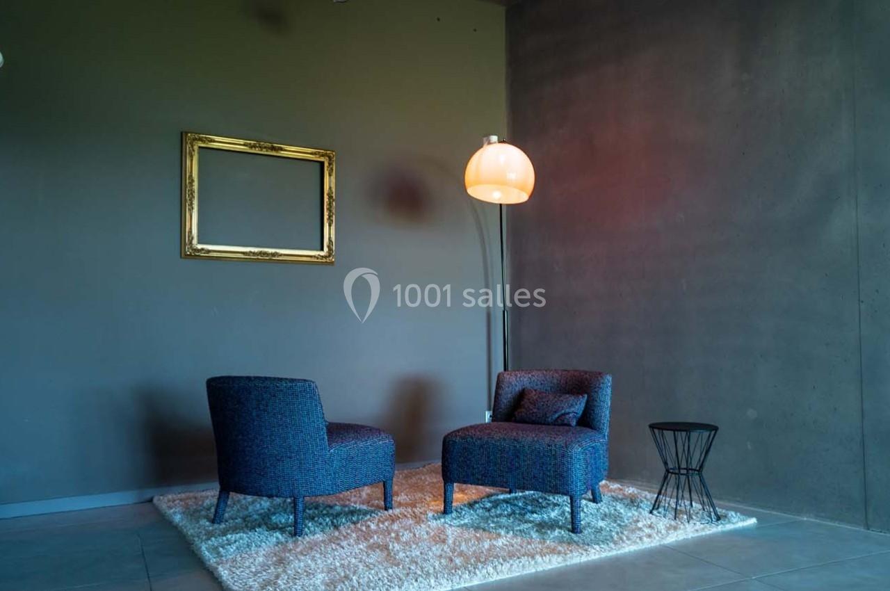 Deux fauteuils bleus sur un tapis blanc, près d'une lampe sur pied et d'un cadre vide, dans une pièce sombre.