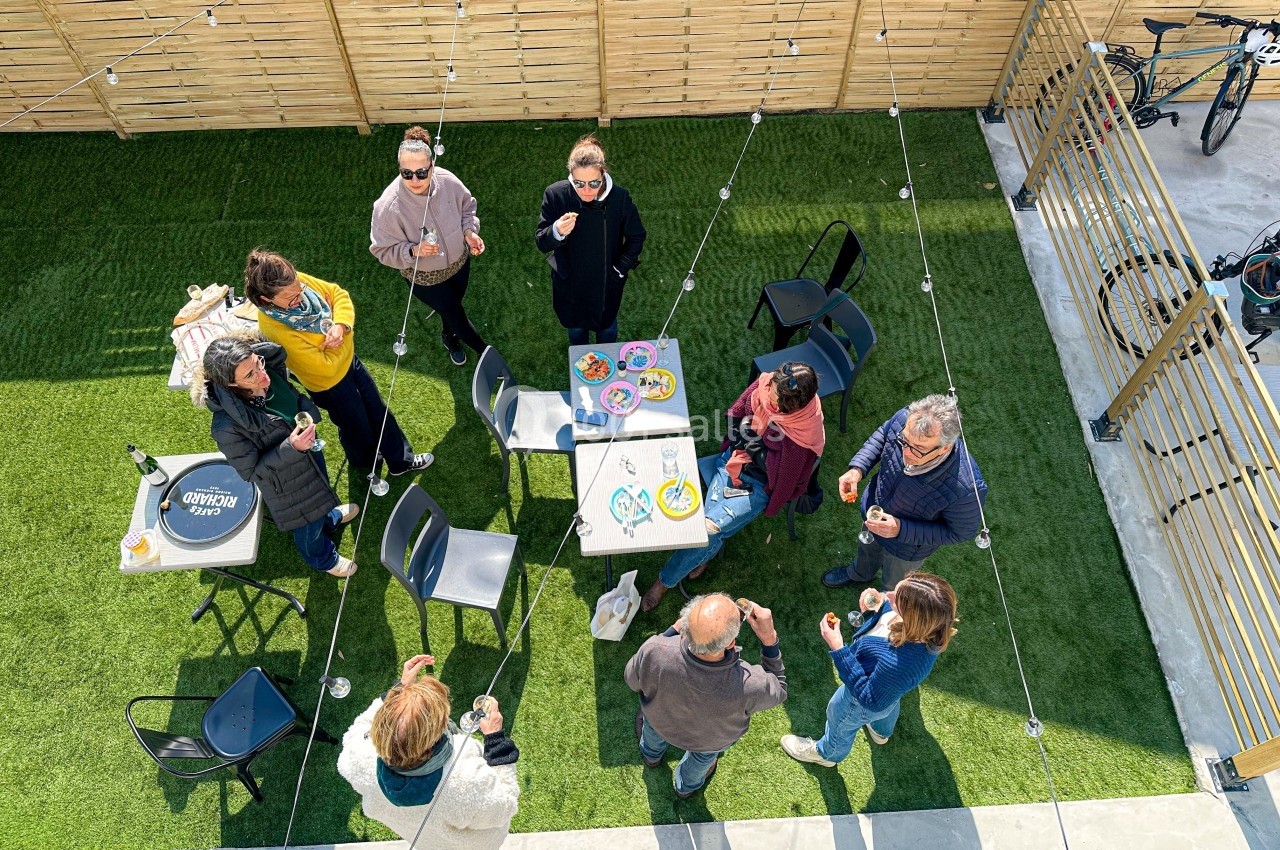 Des personnes debout autour d'une table sur une terrasse avec pelouse artificielle, discutant et partageant un moment…