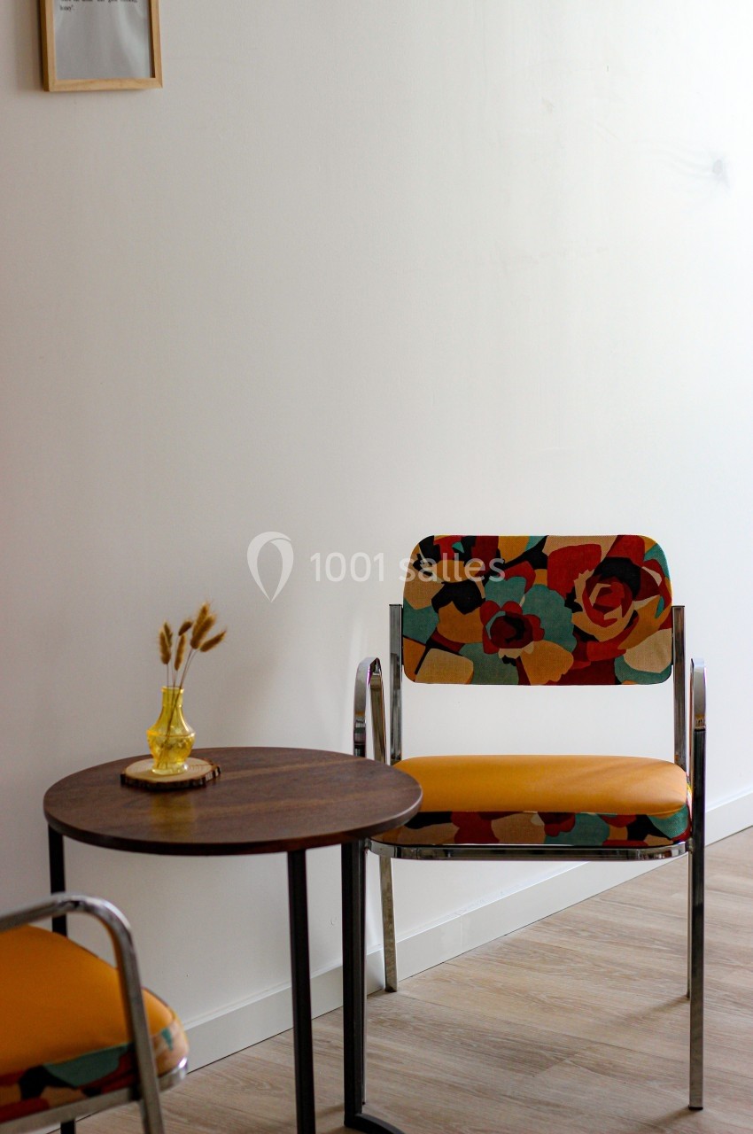 Deux chaises au tissu coloré et une petite table ronde avec un vase jaune, dans une pièce lumineuse.