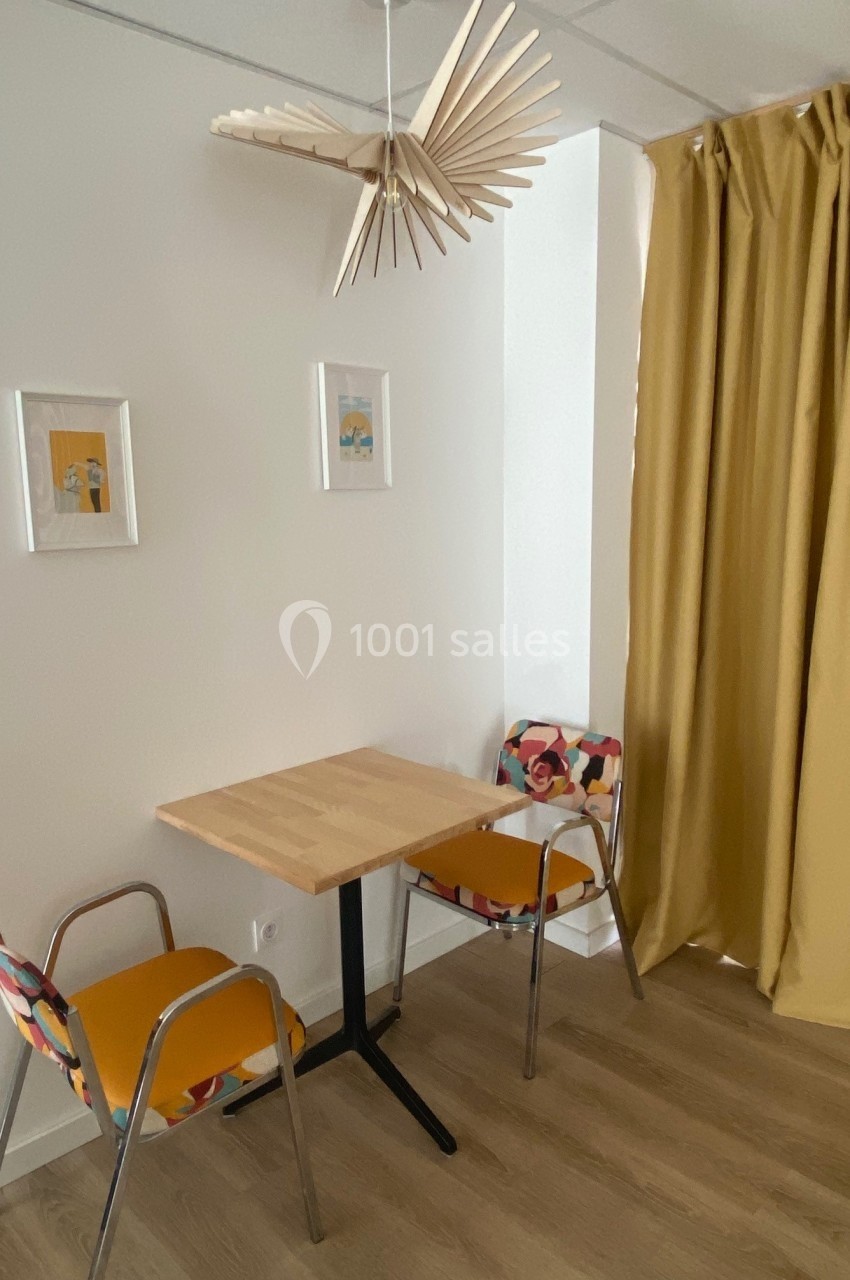 Deux chaises colorées autour d'une table en bois clair, avec des cadres au mur et un rideau jaune en arrière-plan.