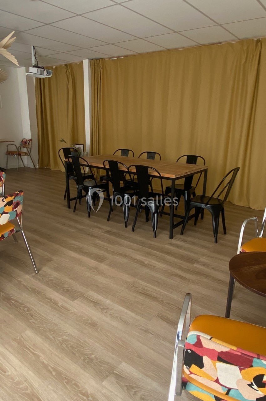Salle lumineuse avec table en bois, chaises noires, rideaux jaunes et fauteuils colorés sur un sol en parquet clair.