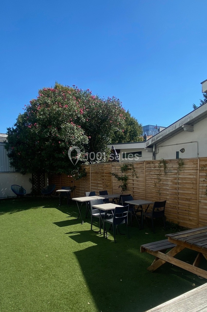 Espace extérieur avec pelouse artificielle, tables, chaises, arbre fleuri et clôture en bois sous un ciel dégagé.