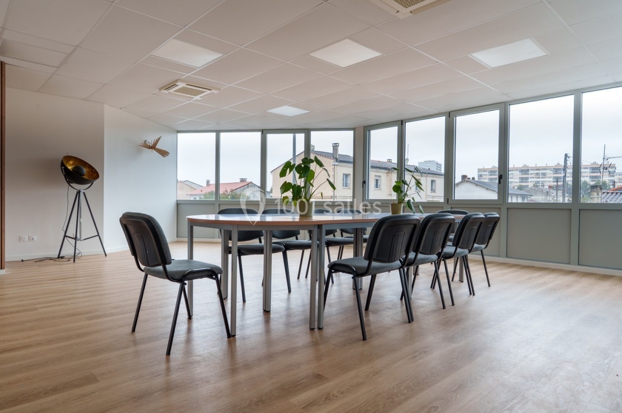 Salle de réunion lumineuse avec table ovale, chaises noires, plantes et grandes fenêtres donnant sur des bâtiments.