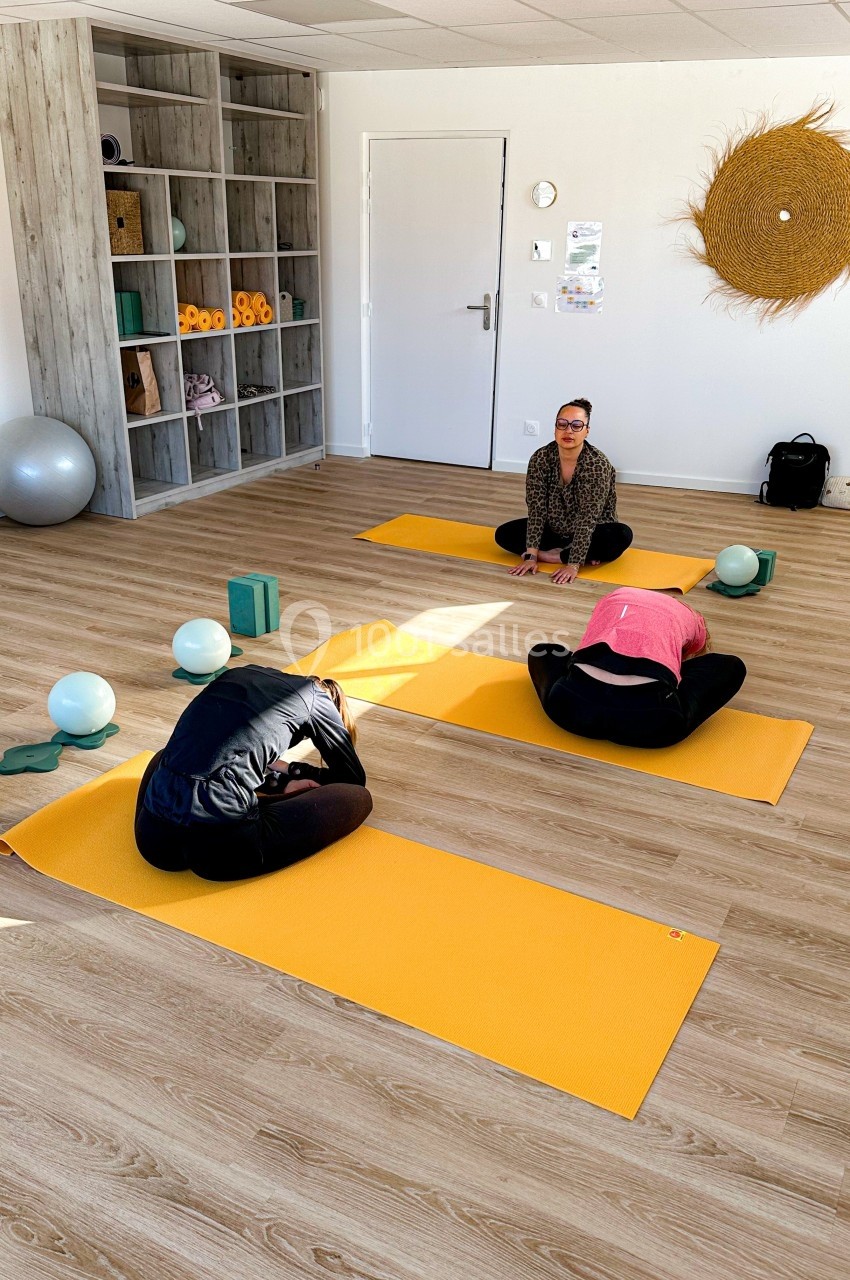 Deux personnes en posture de yoga sur des tapis jaunes dans une salle lumineuse, avec une instructrice les guidant.