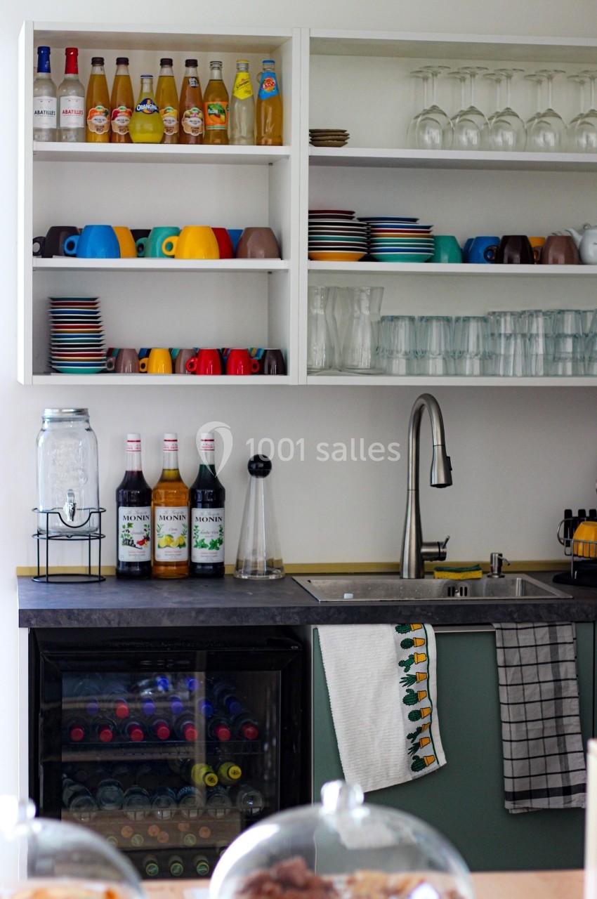 Cuisine avec étagères ouvertes contenant des verres, tasses colorées, bouteilles et un évier en inox à côté d'un mini-frigo.