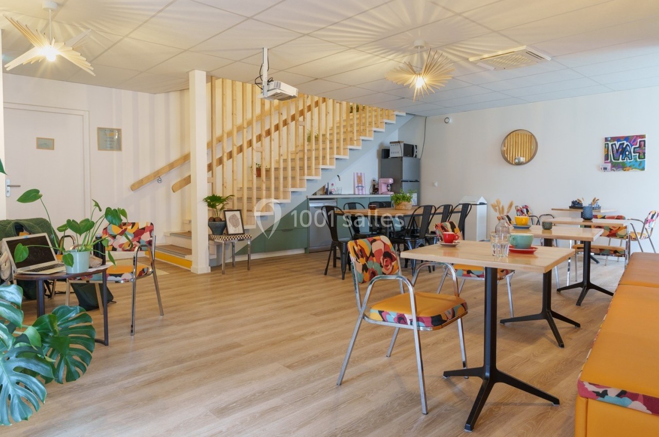 Salle lumineuse avec tables et chaises colorées, escalier en bois, plantes et décoration murale moderne.
