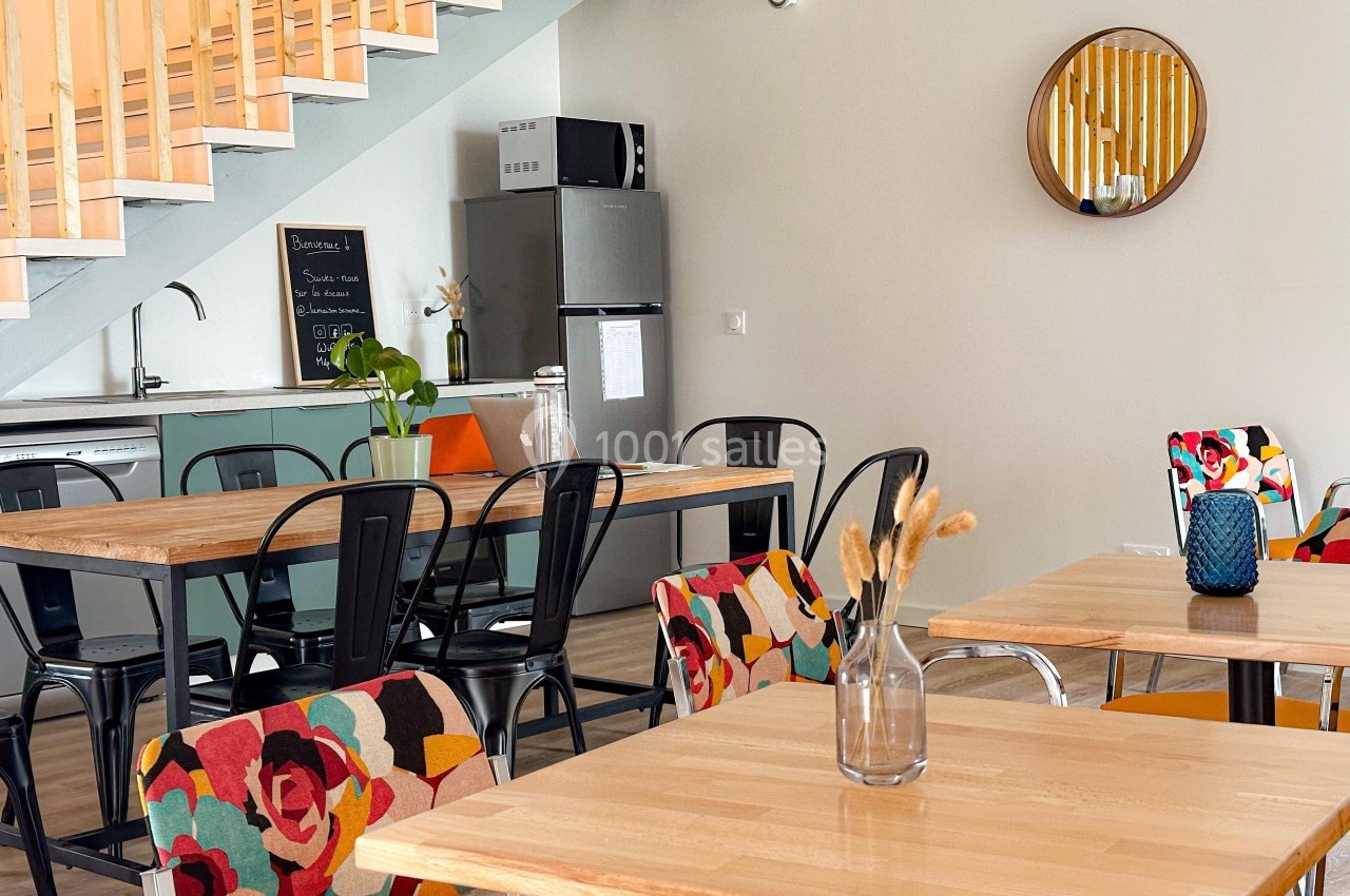 Salle à manger lumineuse avec tables en bois, chaises colorées, cuisine équipée et miroir rond au mur.