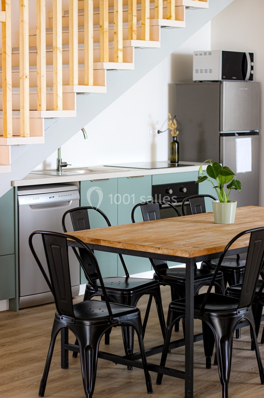 Cuisine moderne avec table en bois, chaises noires, escalier en bois, et électroménagers en arrière-plan.