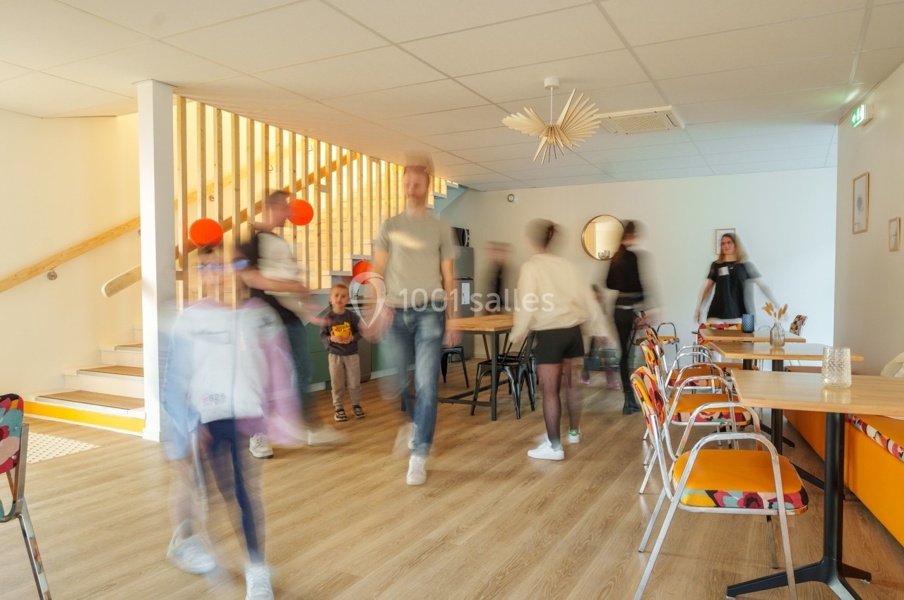 Des personnes floues en mouvement dans une salle lumineuse avec des tables, des chaises colorées et un escalier en bois.