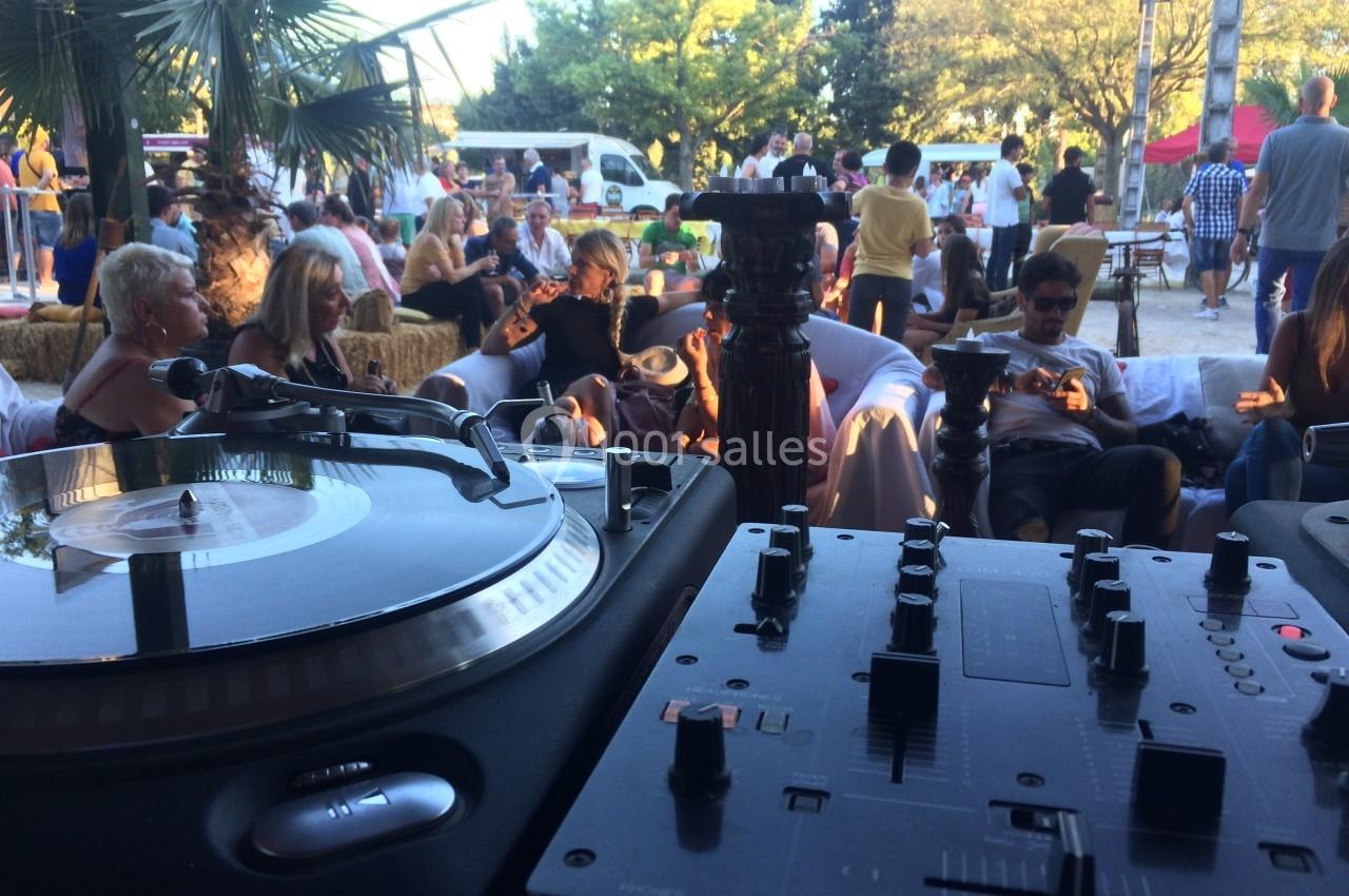 Platines de DJ en premier plan avec des personnes assises en arrière-plan dans un espace extérieur animé.