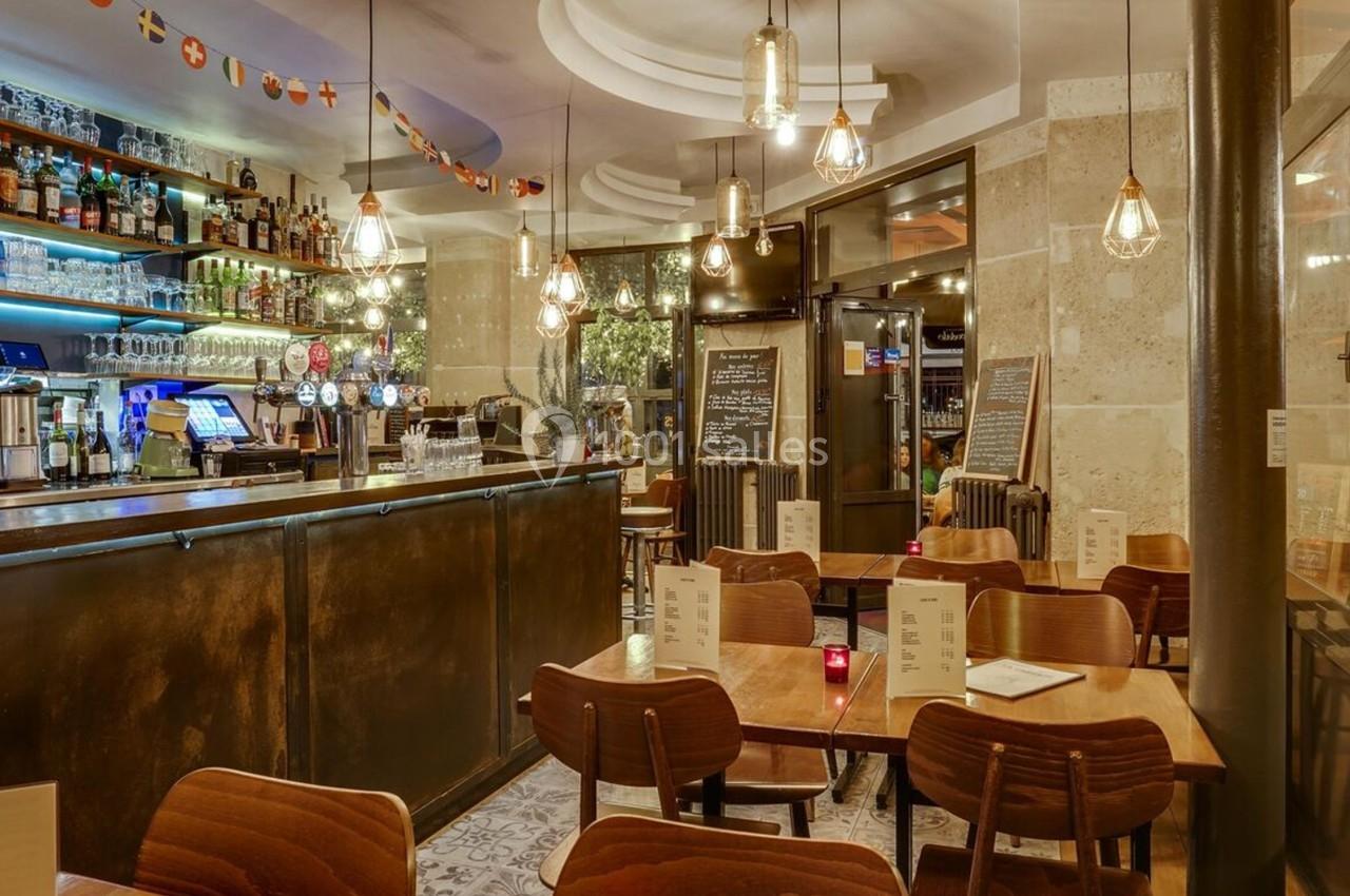 Intérieur d'un café-restaurant avec un bar éclairé, des tables en bois et des suspensions lumineuses.