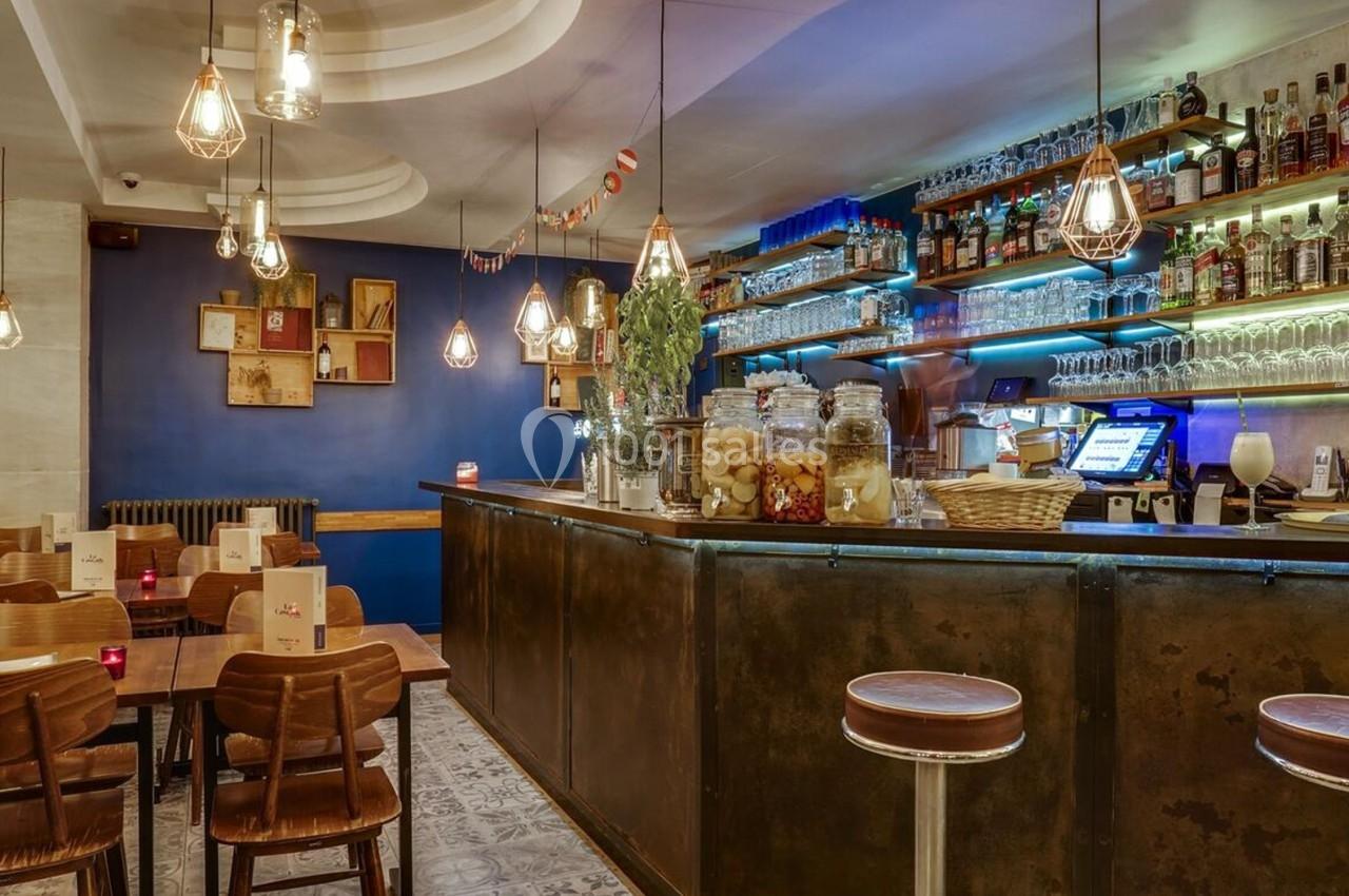 Intérieur d'un bar chaleureux avec comptoir éclairé, étagères de bouteilles et tables en bois.