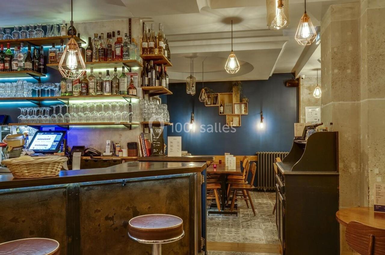 Bar avec comptoir en métal, étagères remplies de bouteilles, éclairage chaleureux et tables en bois dans un décor moderne.