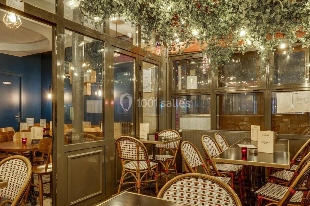 Salle de restaurant avec chaises en rotin, tables en bois et plafond décoré de feuillage et guirlandes lumineuses.